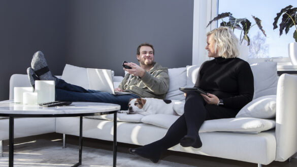 En man och kvinna sitter i en soffa. I mitten av personerna ligger en hund. Mannen har en bärbar dator i knät och håller en tv-fjärrkontroll i vänstra handen. Kvinnan har en Ipad i knät. Båda personerna ler och tittar på varandra.