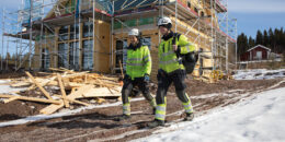 Två leende elmontörer i skyddskläder och hjälm går med utrustning.