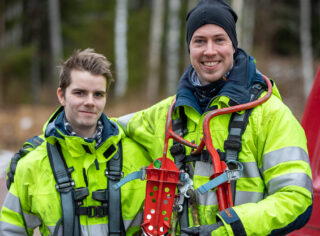 Två elmontörer med skyddskläder och klätterutrustning. Personerna håller om varandra och ler.